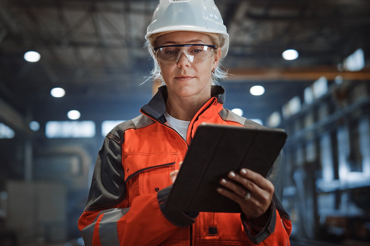 Eine Frau in orangefarbener Jacke und Schutzhelm hält ein Tablet in der Hand